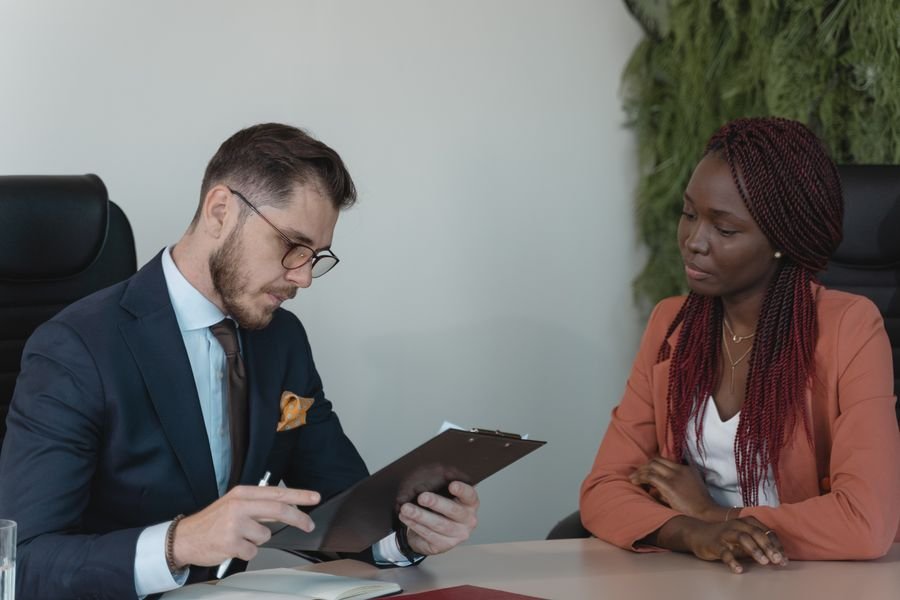 A candidate and an interviewer across a table, one of them reading a CV on a clipboard
